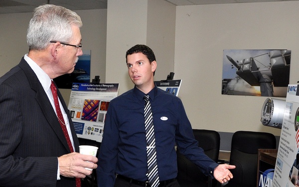 Two men are engaged in a professional presentation setting, with a younger man in a dark shirt and striped tie presenting a research poster to an older man in a suit holding a coffee cup. The poster visible on the right appears to be titled 'Smart Meter Vulnerability Assessment' and is displayed on an easel. The setting appears to be a formal research or academic poster session in an office or conference room environment.