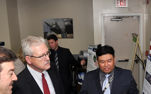 A professional presentation or briefing event is taking place in what appears to be a government or military facility. A man in a dark suit is presenting a research poster board to two other men in business attire, while a fourth person stands in the background. An American flag and an EXIT sign are visible, along with multiple display boards and posters.