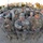 A large formation of U.S. Army soldiers in camouflage uniforms are assembled in what appears to be a staging area near a residential building. The front row of soldiers is equipped with riot control shields and batons, indicating preparation for civil disturbance or crowd control operations. The image is taken with a wide-angle fisheye lens, giving a distorted panoramic perspective of the assembled unit.