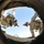 The image is taken from inside a large concrete culvert or pipe looking upward, capturing two soldiers in U.S. Army combat uniforms peering down into the opening. The fisheye perspective creates a circular framing effect with the pipe's interior edge forming a dark border around the scene. The soldiers appear to be inspecting or searching the structure against a clear blue sky backdrop.