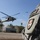 A close-up shot of a U.S. Army soldier in ACU (Army Combat Uniform) digital camouflage, with a unit patch and 'MP' (Military Police) tab visible on the shoulder. In the background, a UH-60 Black Hawk helicopter is hovering or landing on what appears to be a landing zone, with a second helicopter visible in the far distance. The image uses a shallow depth of field, keeping the uniform patch in sharp focus while the background remains blurred.