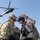 Two military personnel in camouflage uniforms are engaged in a briefing or coordination discussion in the foreground, with one pointing in a direction. A UH-60 Black Hawk helicopter is visible in flight in the background against a clear blue sky. The scene appears to depict an active military operation or training exercise.