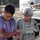 A military training exercise or disaster response drill is depicted, showing a soldier named Sanchez (visible on name tape) assisting a simulated casualty with moulage (theatrical makeup) wounds. The scene includes a staged disaster environment with a damaged vehicle, debris, and multiple military personnel in ACU (Army Combat Uniform) pattern. The setting appears to be a controlled training scenario rather than an actual emergency.