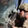 A military soldier in MultiCam/OCP camouflage uniform and helmet is examining or clearing shattered glass from a vehicle window. The glass is extensively fractured in a spiderweb pattern consistent with impact damage. The scene appears to be part of a rescue operation, training exercise, or post-incident assessment.