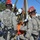 Military personnel in U.S. Army ACU (Army Combat Uniform) camouflage are conducting a search and rescue training exercise. They are using a tripod hoist system to extract a simulated casualty secured in an orange rescue litter/basket. Personnel are equipped with gas masks labeled 'TRNG' (training) and various rescue equipment.