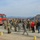 A large group of military personnel in camouflage uniforms are gathered on a gravel area in a desert/mountainous environment. A red Los Angeles Fire Department engine (Unit 143) is parked to the left. The gathering appears to be a briefing, exercise, or coordination event involving both military and civilian personnel.
