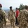 A group of military personnel in camouflage uniforms are gathered outdoors on elevated terrain with mountains visible in the background. A central figure wearing a green beret appears to be briefing or conversing with the group. At least one civilian in a red jacket is also present among the military personnel.