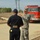 A U.S. Army soldier in ACU (Army Combat Uniform) camouflage is speaking with a civilian emergency responder or fire department official wearing a dark navy uniform and cap. They appear to be coordinating at what looks like a demolition or disaster site, with rubble visible to the left and a red fire engine parked nearby. The setting suggests an inter-agency emergency response or training exercise.