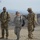 Three military personnel are standing outdoors in an arid, mountainous environment. Two individuals appear to be wearing U.S. military uniforms (OCP/MultiCam and ABU patterns), while the third wears a different camouflage pattern consistent with an African nation's military. They appear to be engaged in conversation, with one person gesturing.