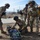 Military or special operations personnel are conducting what appears to be a training exercise at an outdoor facility. Several personnel in multicam/OCP camouflage uniforms with tactical gear and communications headsets are working with a stretcher/litter on the ground. One individual is gesturing directionally while others prepare the medical evacuation equipment.
