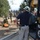 The image shows a multi-agency emergency response or training exercise involving Los Angeles County Fire Department personnel and military soldiers. A firefighter in a white helmet and navy shirt stands near a Los Angeles County Fire vehicle marked 'TL1255C', observing soldiers working with ropes and rescue equipment on the ground. The scene appears to be an outdoor training or disaster response operation in a dry, semi-arid environment.