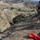 A military or tactical personnel in full combat gear stands at the edge of a steep cliff or ridgeline, overseeing a rope/chain rigging operation in an arid, hilly terrain. A second individual is visible lower on the slope, apparently secured by the rigging system. The setting appears to be a training exercise or field operation in a dry, mountainous region with a small facility visible in the valley below.
