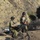 Two personnel in military/tactical camouflage uniforms and helmets are carrying a folded litter/stretcher in a rugged desert canyon environment. A crashed or disabled vehicle is visible in the background at the base of a steep sandy hillside. A rope or cable system is visible in the foreground, suggesting a rope rescue or recovery operation is underway.