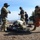 Military or special operations personnel in multicam/camouflage uniforms and flight helmets are conducting a medical evacuation exercise on a hilltop in an arid, mountainous environment. A simulated casualty is secured on a Stokes litter/basket stretcher while team members attend to the patient and manage equipment. Observers in civilian and fire department attire watch from the background, suggesting this is a joint training exercise.