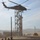 A military helicopter, appearing to be a UH-60 Black Hawk, hovers low over a training facility in an arid, dusty environment. Personnel can be seen on the ground and what appears to be a soldier rappelling or being inserted from the helicopter near a tall metal training tower. The scene suggests a military fast-rope or rappel training exercise at a dedicated training installation.
