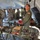Military personnel in various uniforms are gathered inside a large field medical tent during what appears to be a training exercise or drill. Multiple mannequins or simulated casualties are positioned on military stretchers/litters, with a prominent figure in an olive flight suit wearing a US flag patch standing near them. The setting suggests a mass casualty triage or field hospital exercise.