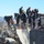A team of urban search and rescue (USAR) personnel are working atop a collapsed concrete structure surrounded by rubble. The responders are equipped with helmets, protective gear, and power tools including what appears to be a concrete-cutting chainsaw or rotary saw. The scene appears to be either an active disaster response or a training exercise set against a mountainous backdrop.