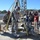 Military personnel in U.S. Army combat uniforms are conducting a confined space rescue training exercise, using a tripod rigging system over what appears to be a manhole or underground access point. Several soldiers wearing red and white safety helmets are actively working the rescue equipment while observers and media personnel with cameras document the exercise. The setting appears to be a military training facility with hills and infrastructure visible in the background.