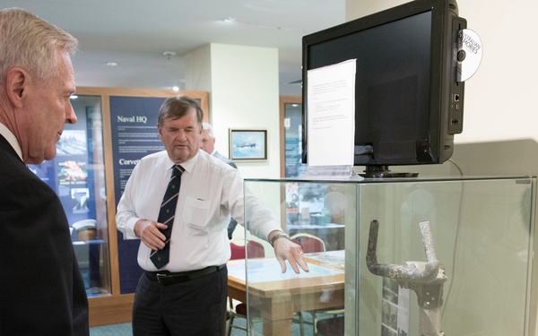 Two men in formal attire are examining a museum or exhibition display case containing what appears to be a corroded metal artifact, likely of naval or historical significance. A man in a white shirt and tie is gesturing toward the glass display case, appearing to explain or present the artifact to the other man in a dark suit. The setting appears to be a naval heritage or military museum, evidenced by display panels in the background labeled 'Naval HQ' and 'Corvette'.