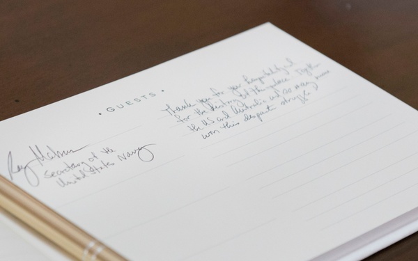 The image shows an open guest book on a wooden surface with a gold and silver pen resting across it. A signature and handwritten message are visible on the right-hand page, with the signer identifying themselves as 'Secretary of the United States Navy.' The handwritten note appears to reference the US and Australia working together.