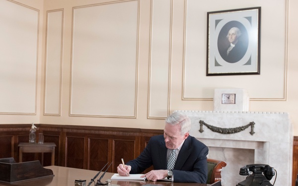 An older man in a dark suit and tie is seated at a large wooden desk, writing on paper in what appears to be a formal government or judicial office. The room features wood paneling, a marble fireplace, and a framed portrait on the wall that resembles George Washington. The setting evokes a historic or official government workspace with period-appropriate furnishings.