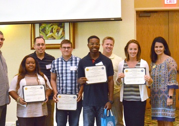 A group of nine people pose for a photo in what appears to be a banquet or conference room. Four individuals are holding certificates or awards, suggesting this is a recognition or awards ceremony. The setting includes patterned carpet, framed artwork, and an exit door visible in the background.