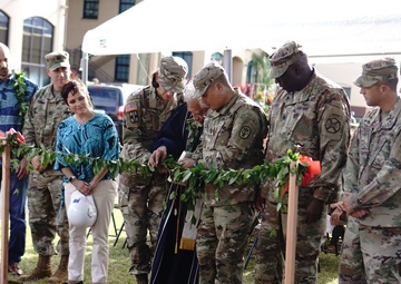 A military groundbreaking ceremony is taking place outdoors, likely at a U.S. Army installation in Hawaii, evidenced by the lei garlands and tropical vegetation. Multiple soldiers in Army Combat Uniforms (ACU/OCP pattern) are gathered around a freshly turned dirt plot, with golden ceremonial shovels visible in the foreground. A chaplain or officiant in dark robes with gold stole appears to be leading a blessing or prayer.