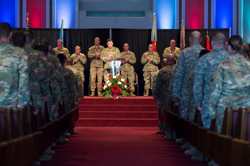 A military ceremony is taking place in what appears to be a chapel or auditorium, with one soldier speaking at a podium on a raised stage while others stand in formation on either side of a center aisle. A floral arrangement of red and white flowers is placed at the base of the stage steps. The venue is lit with red and blue lighting, and multiple military flags are visible on stage.