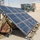 A portable military solar panel array is deployed on sandy ground at what appears to be a military installation or base. The solar panels show significant wear and damage, suggesting heavy field use. Military vehicles and a building with solar panels are visible in the background.