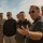 A group of individuals, including at least two uniformed military personnel in desert camouflage and several civilians, are gathered outdoors in an arid, desert-like environment. A man in the foreground wearing sunglasses, a dark polo shirt, and a lanyard reading 'Colorado' is speaking and gesturing with his hand. The setting suggests a military installation visit or briefing in a desert location.
