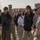 A military briefing or orientation is taking place outdoors at what appears to be a military installation or training facility. A Marine in desert camouflage (MARPAT) uniform is addressing a mixed group of military personnel and civilians. The setting appears arid with sandy ground and temporary structures visible in the background.