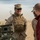 Two U.S. Marines in desert camouflage uniforms and tactical gear are engaged in conversation with a civilian wearing a red sweatshirt in an arid, desert environment. The central Marine is smiling and wearing sunglasses, a patrol cap, and a plate carrier vest with visible pouches and a radio. Military equipment, likely an artillery piece or weapons system, is visible on the left side of the frame.