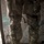 Close-up photograph of a heavily equipped military or special operations personnel from the waist down, showing detailed tactical gear on both legs. The individual is wearing multicam/OCP camouflage pattern clothing with extensive leg-mounted equipment including thigh holsters and knee protection. The setting appears to be an indoor or semi-sheltered position with camouflage netting visible in the background.