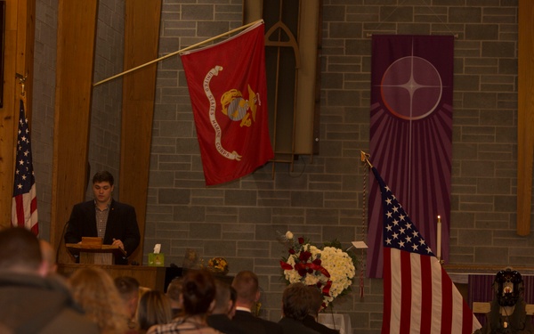 A military memorial or funeral service is taking place inside a church or chapel. A young man in a dark blazer is speaking at a podium while attendees sit in wooden pews. The altar area displays a U.S. Marine Corps flag, an American flag, a floral wreath, military gear, and what appear to be photographs of the deceased.