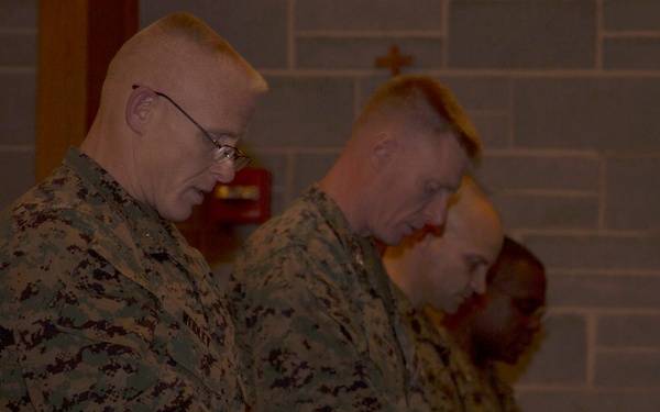 Three individuals in U.S. Marine Corps MARPAT digital camouflage uniforms are standing with heads bowed, appearing to be in prayer or a moment of reflection. They are holding what appear to be papers or cards, and a cross is visible on the wall behind them, suggesting a chapel or religious setting. The lighting is dim, creating a subdued atmosphere consistent with a religious service.