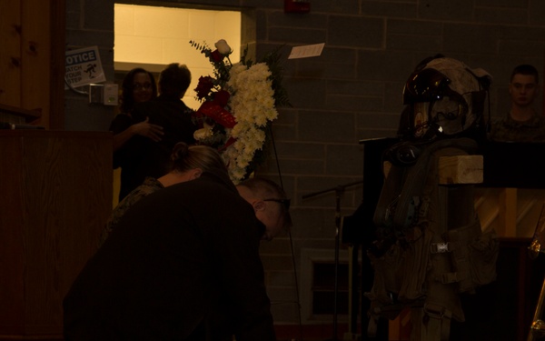 A dimly lit memorial ceremony appears to be taking place in what looks like a military or community hall. A person is kneeling on the floor near a display that includes a military EOD (Explosive Ordnance Disposal) bomb suit, photographs on an easel, and a floral arrangement, suggesting this is a memorial service for a fallen service member. An American flag is visible in the background along with other attendees.