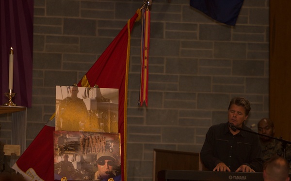 A solemn military memorial or memorial service is taking place in what appears to be a chapel or auditorium. A musician is playing a Yamaha keyboard at a podium while an easel displays photographs of what appears to be the deceased service member. Military flags and personnel in uniform are visible among the gathered attendees.
