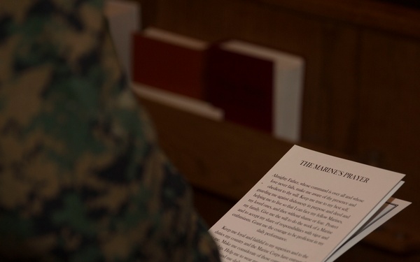 A person wearing U.S. Marine Corps MARPAT camouflage uniform is holding a printed document titled 'THE MARINE'S PRAYER' in what appears to be a chapel or religious setting with wooden pews visible in the background. The document contains multiple paragraphs of prayer text. The setting and document suggest a religious or ceremonial military event.