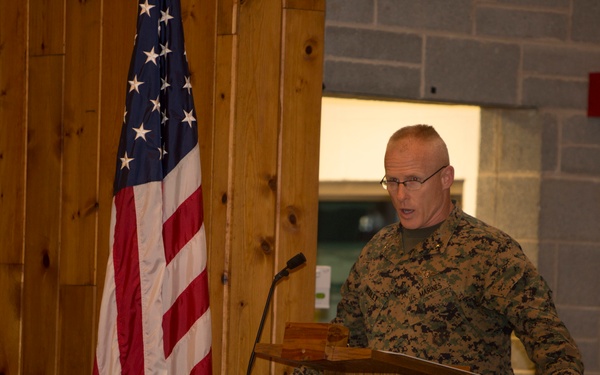 A U.S. Marine in woodland MARPAT camouflage uniform is speaking at a wooden podium during what appears to be an official military briefing or ceremony. An American flag with gold fringe stands to his left, and the setting appears to be an indoor military facility with wood-paneled walls and cinder block construction. The speaker is wearing glasses and appears to be addressing an audience.