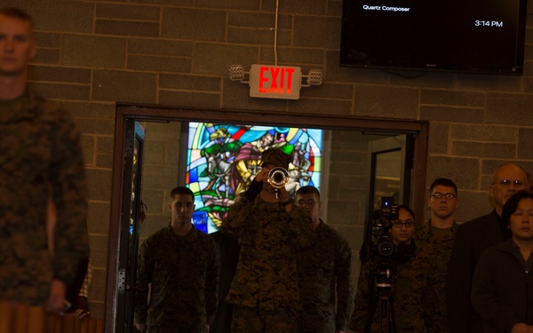 A military ceremony is taking place inside what appears to be a chapel or church, identifiable by wooden pews and a stained glass window visible through the doorway. A service member in camouflage uniform is playing a brass instrument (likely a bugle or trumpet) at the entrance aisle, while other uniformed personnel and civilians stand nearby. A video camera on a tripod is present, suggesting the event is being recorded.