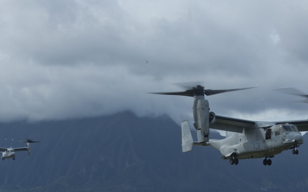 Two military tiltrotor aircraft, identified as Bell Boeing V-22 Ospreys, are captured in flight over what appears to be a military installation near a coastal area with a dramatic mountainous backdrop. The foreground shows military base buildings, while a large body of water and steep green mountains shrouded in clouds are visible in the background. The setting is consistent with a Hawaiian military base, likely Marine Corps Base Hawaii (Kaneohe Bay).