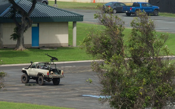 The image depicts what appears to be a military training exercise taking place on or near a military installation. Several soldiers in combat gear are positioned in prone and crouching positions on a grassy area, appearing to engage a white pickup truck mounted with what appears to be a weapon system. The setting includes parking lots, buildings, and civilian vehicles in the background, consistent with a base or installation environment.