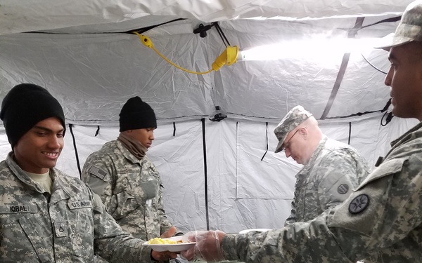 U.S. Army soldiers are participating in a field mess operation inside a military tent. A soldier identified by the name tape 'IQBAL' is being served food from insulated green military food containers. The setting appears to be a forward operating or field training environment based on the temporary tent structure and field equipment.