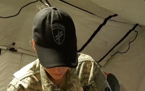 A U.S. Army soldier wearing ACU (Army Combat Uniform) with the name tape 'YERDON' is handling MRE (Meal, Ready-to-Eat) packages inside a military tent. The soldier is wearing a black Airborne cap and is sorting or distributing a large quantity of MRE pouches from an open cardboard box. The setting appears to be a field operation or training exercise.