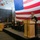 A formal U.S. Navy military ceremony, likely a change of command or commissioning, is taking place aboard a naval vessel in what appears to be a hangar bay. A naval officer in dress blues stands at a podium speaking, while several other uniformed officers are seated on a raised stage behind him. A large American flag serves as a prominent backdrop, along with what appears to be a state flag on the right.