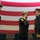 Three U.S. Navy officers in dress uniforms are participating in a formal military ceremony in front of a large American flag. The central officer is rendering a salute, suggesting this may be a change of command, retirement, or similar official military ceremony. The setting appears to be an indoor venue such as a hangar or gymnasium.