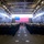 A formal U.S. Navy military ceremony is taking place inside the well deck or hangar bay of a large naval vessel. Rows of sailors in dress uniforms are standing at attention facing a podium and a large American flag displayed at the far end of the compartment. The setting, formation, and flag display are consistent with a commissioning, change of command, or similar official naval ceremony.