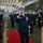 A formal U.S. Navy military ceremony is taking place inside what appears to be a ship's hangar bay. A senior officer wearing a white-topped combination cover and multiple service ribbons walks along a red carpet while personnel on both sides render salutes. The setting and decorations suggest this is a change of command or commissioning ceremony.