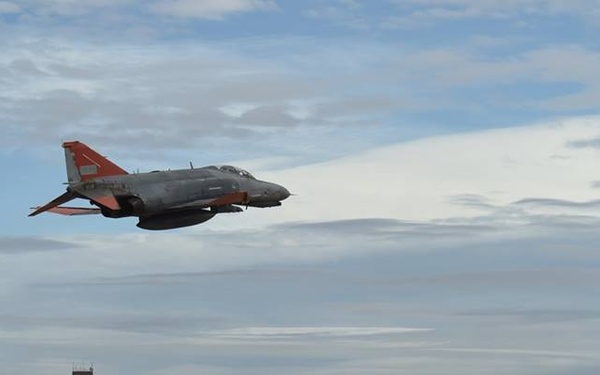 A military jet aircraft, identifiable as an F-4 Phantom II painted in gray with distinctive orange markings on the tail and wings, is captured in low-level flight over a desert airfield. The aircraft appears to be configured as a QF-4 drone or target aircraft based on its orange high-visibility markings. An air traffic control tower and sparse airfield infrastructure are visible in the background against a mountain range.