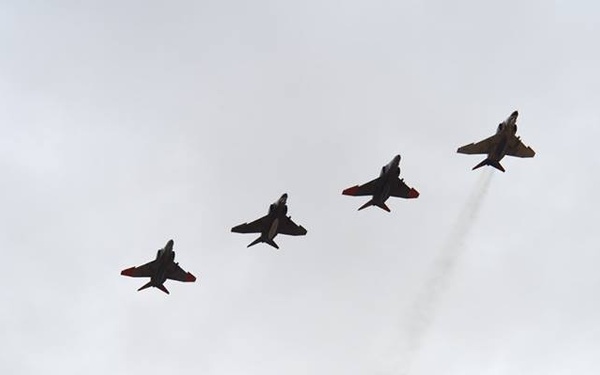 Four jet aircraft are flying in a diagonal line formation against an overcast sky. The aircraft display red and dark coloring consistent with aerobatic display teams, and at least one aircraft is emitting a white smoke trail. The formation appears to be climbing or banking in a coordinated maneuver.
